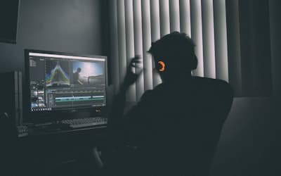 A person in silhouette wearing headphones editing video on a computer in a dark room